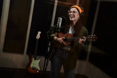 Ukulele çalan ve profesyonel bir stüdyoda şarkı kaydederken mikrofona şarkı söyleyen genç bir kadın ve kadın sanatçının portresi