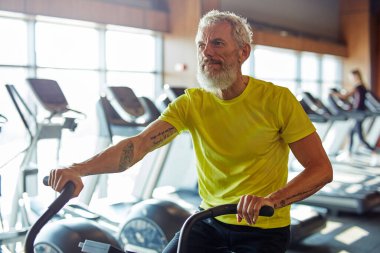 Formda kalmak. Spor kıyafetli olgun bir sporcu spor salonunda bisiklet sürüyor.