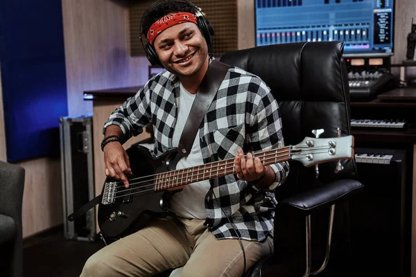 Retrato de jovem feliz, artista masculino em fones de ouvido sorrindo de lado, tocando guitarra enquanto sentado no estúdio de gravação — Fotografia de Stock