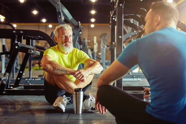 Vilar efter träningen. Atletisk medelålders man diskuterar något med fitness instruktör eller personlig tränare när du sitter tillsammans på golvet på gym — Stockfoto