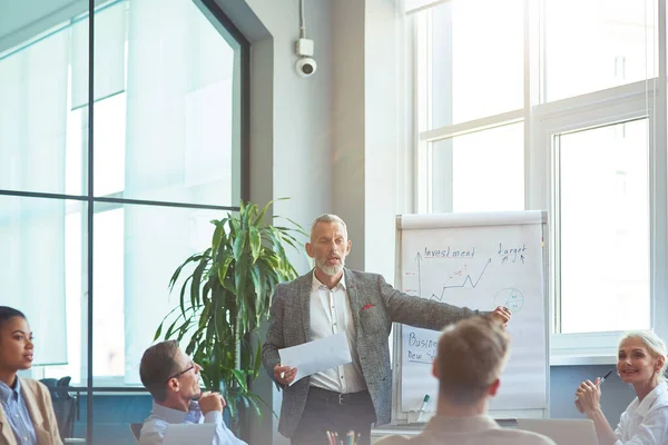 Homme d'affaires mature confiant pointant du doigt le tableau blanc et expliquant quelque chose à ses collègues tout en ayant une réunion dans le bureau moderne. Bussiness personnes travaillant ensemble — Photo