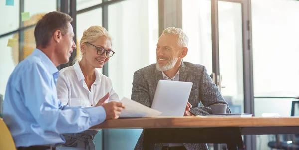 Gente de negocios exitosa trabajando juntos, tres compañeros de trabajo felices interactuando, discutiendo algo y sonriendo mientras están sentados en el escritorio en la oficina moderna o en el espacio de coworking —  Fotos de Stock