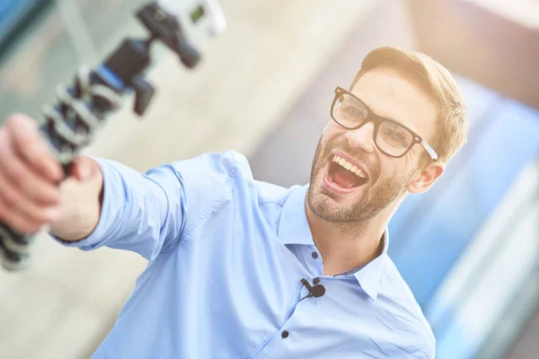 Criar novos conteúdos. Jovem blogueiro feliz do sexo masculino vestindo camisa azul e óculos segurando um gimbal com smartphone e gravação de vídeo para seu blog enquanto está de pé ao ar livre — Fotografia de Stock