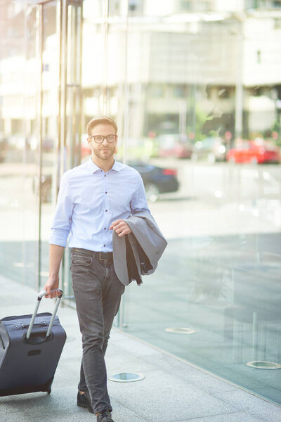 Иду к терминалу аэропорта. Вертикальный снимок молодого уверенного бизнесмена, идущего по улицам города и вытаскивающего свой чемодан