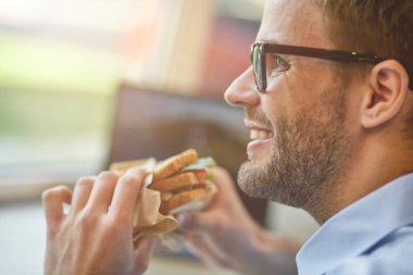 Öğle yemeği vakti. Kafede uzaktan çalışırken sandviç yiyen mutlu bir gencin yakın çekimleri.