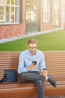 İş e-postası yazıyorum. Yakışıklı bir iş adamının dışarıda bankta otururken akıllı telefon kullanıp kahve içtiği dikey çekim.