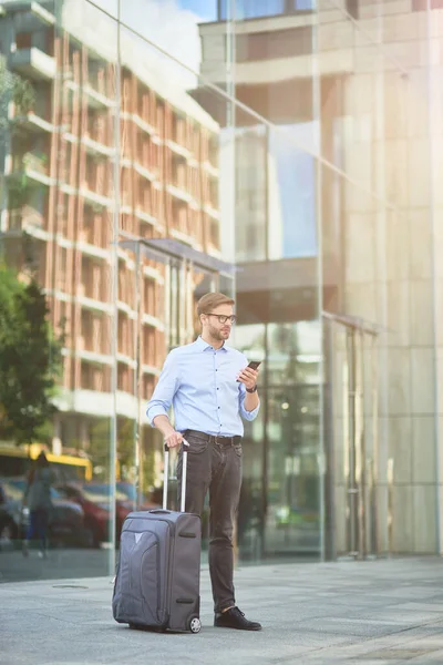 Pionowe ujęcie eleganckiego przystojnego biznesmena z walizką za pomocą smartfona i odwracając wzrok stojąc na zewnątrz, czekając na taksówkę — Zdjęcie stockowe
