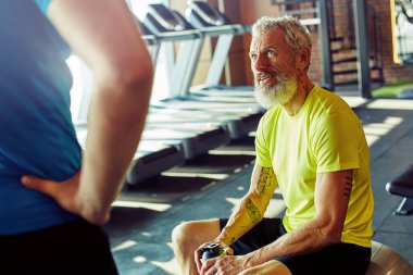 Bitkin hissediyorum. Spor giyim kuşağında orta yaşlı bir adam elinde bir şişe su tutarken spor salonunda ya da spor kulübünde antrenörle konuşurken kişisel antrenörle konuşuyor.