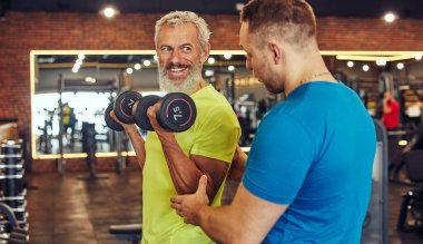 Spor salonunda beden eğitimi hocasıyla çalışıyorum. Spor giyim sektöründe genç profesyonel antrenörün yardımıyla ağırlık egzersizi yapan olgun bir adam.