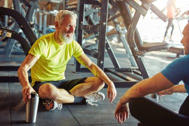 Kişisel antrenörle konuşuyorum. Orta yaşlı mutlu bir adam beden eğitimi hocasıyla tartışıyor ve spor salonunda birlikte otururken gülümsüyor.