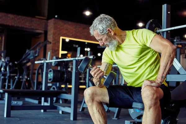 40 'ından sonra kasların gelişiyor. Spor kıyafetli güçlü bir adam spor salonunda spor yaparken ağır dambıllar kaldırıyor ve pazılarını pompalıyor. — Stok fotoğraf