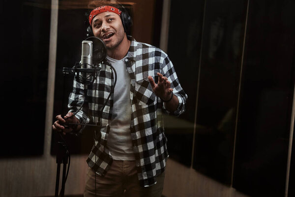 Portrait of young man, male artist looking focused, singing into a condenser microphone while recording a song in a professional studio