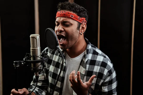 Portrait of young man, hip hop artist singing with closed eyes into a condenser microphone while recording a song in a professional studio
