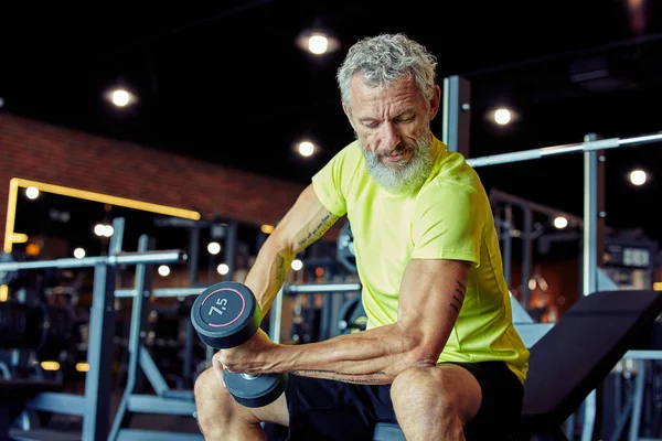 Ağırlık çalışması. Spor kıyafetleriyle odaklanmış, ağırlık kaldıran ve spor salonunda antrenman yaparken bisepslerini pompalayan olgun bir adam.