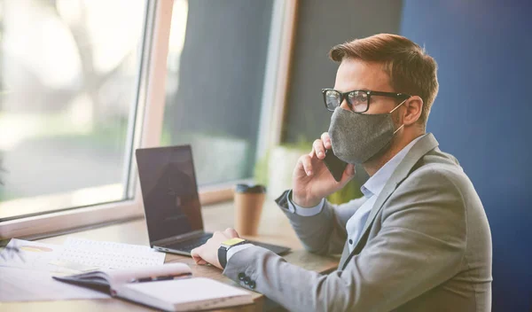 Vista lateral de un hombre de negocios ocupado que usa mascarilla protectora usando computadora portátil y hablando por teléfono con su pareja mientras trabaja remotamente en la cafetería durante el brote de covid 19 — Foto de Stock