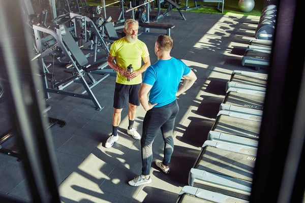 Tiro completo de um homem de meia-idade em sportswear falando com instrutor de fitness ou personal trainer enquanto exercita-se no ginásio ou clube esportivo — Fotografia de Stock