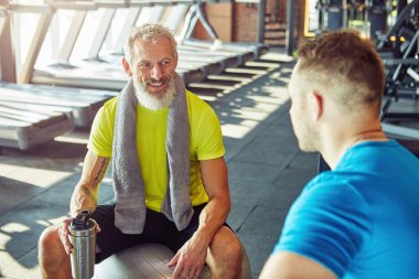 Ara veriyoruz. Spor kıyafetli, elinde su şişesi olan orta yaşlı bir adam spor salonunda ya da spor kulübünde fitness hocasıyla konuşuyor.