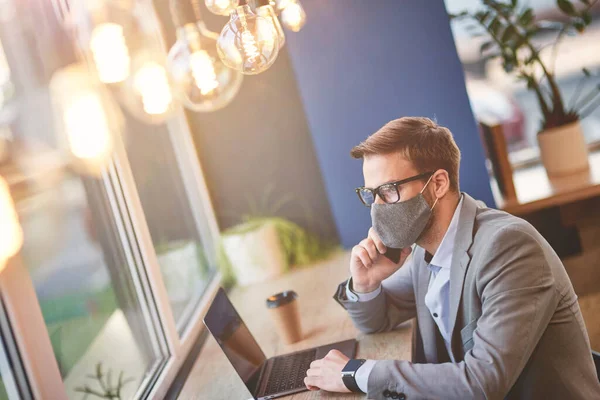 Joven hombre de negocios que usa mascarilla protectora usando computadora portátil y hablando por teléfono con su pareja mientras trabaja remotamente en la cafetería durante el brote de covid 19 — Foto de Stock