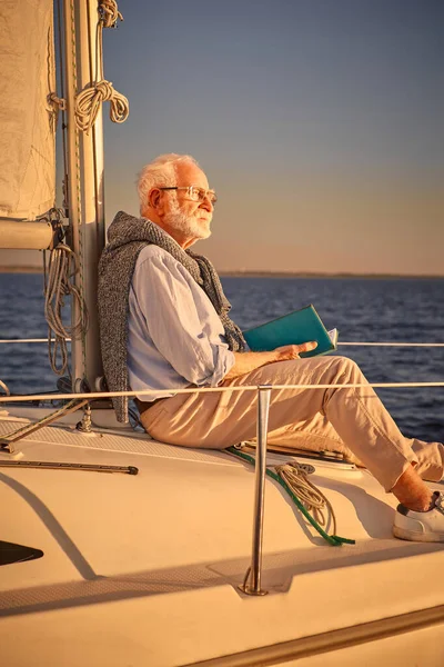 Plan vertical d'un homme retraité détendu lisant un livre et profitant d'un coucher de soleil incroyable assis sur le côté de son voilier ou pont de yacht flottant dans la mer bleue calme — Photo