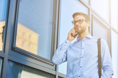 Mavi gömlekli yakışıklı beyaz iş adamı dışarıda dikilirken telefonla konuşuyor.