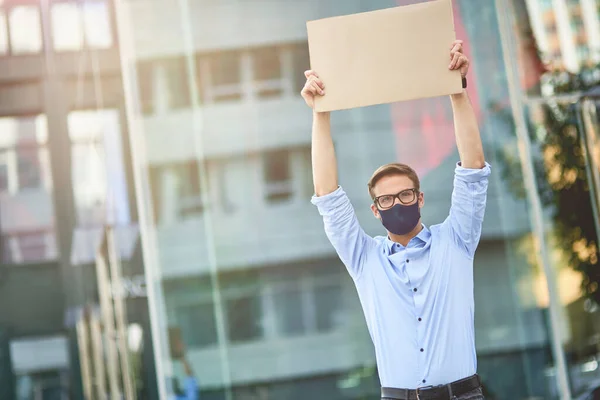 Joven hombre caucásico, activista masculino con máscara protectora negra sosteniendo un cartel vacío sobre la cabeza mientras está de pie en la calle de la ciudad. Protesta durante el brote de coronavirus — Foto de Stock