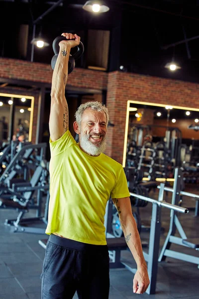 Vertical tiro de um homem caucasiano maduro sorridente em sportswear exercício com halteres no ginásio — Fotografia de Stock