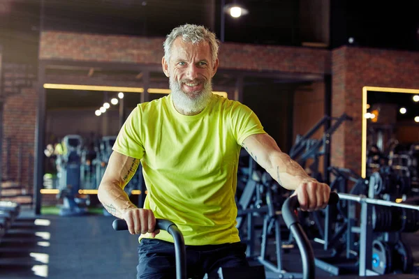 Retrato de homem atlético maduro exausto em sportswear fazendo ciclismo em bicicletas de exercício no ginásio — Fotografia de Stock