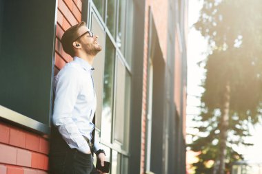 Genç ve mutlu bir işadamının kablosuz kulaklık ve cep telefonuyla dışarıda duvara yaslanmış müzik dinlemesinin yan görüntüsü.