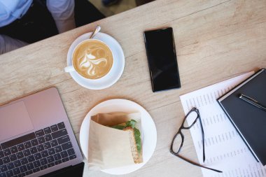 Kahvaltı yapıyorum. Kahve kapuçino, sandviç, dizüstü bilgisayar ve akıllı telefon kafedeki ahşap masada.