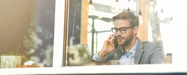 Joven hombre de negocios guapo con anteojos hablando por teléfono mientras trabaja remotamente en la cafetería — Foto de Stock