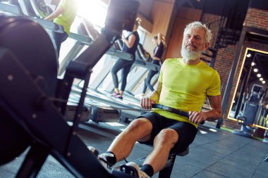 Formda ve sağlıklı kalmak. Spor giyimli güçlü olgun bir sporcu spor salonunda kürek çekme makinesinde egzersiz yapıyor.