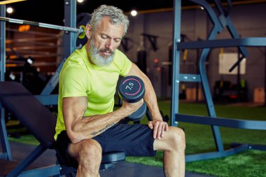 Her yaşta vücut geliştirme. Spor kıyafetli güçlü bir adam spor salonunda spor yaparken ağır dambıllar kaldırıyor ve pazılarını pompalıyor.
