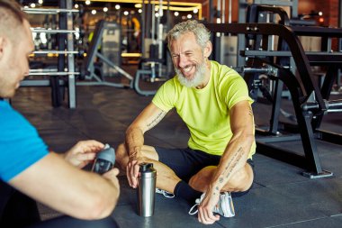 Kişisel antrenörle egzersiz yapıyorum. Orta yaşlı mutlu bir adam beden eğitimi hocasıyla tartışıyor ve spor salonunda birlikte otururken gülümsüyor.