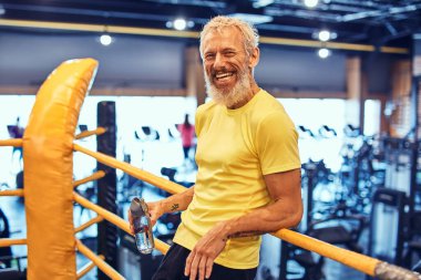 Spor kıyafetli neşeli, olgun bir adam kameraya bakıyor ve gülümsüyor, ringde duruyor ve spor salonundaki boks antrenmanından sonra dinleniyor.
