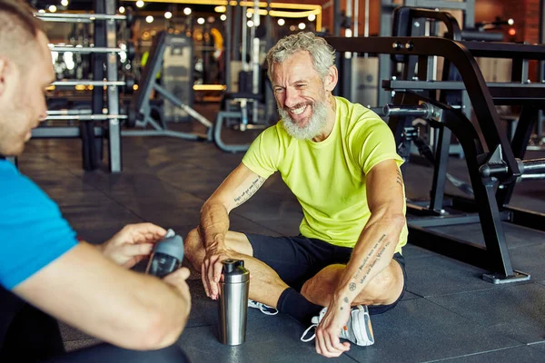 Exercício com personal trainer. Homem de meia-idade feliz discutindo algo com instrutor de fitness e sorrindo enquanto sentados juntos no chão no ginásio — Fotografia de Stock