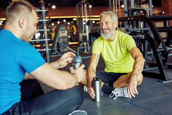 Snyggt snack efter träningen. Glada medelålders man i sportkläder hålla flaska vatten och prata med fitness instruktör när du sitter tillsammans på golvet på gym — Stockfoto