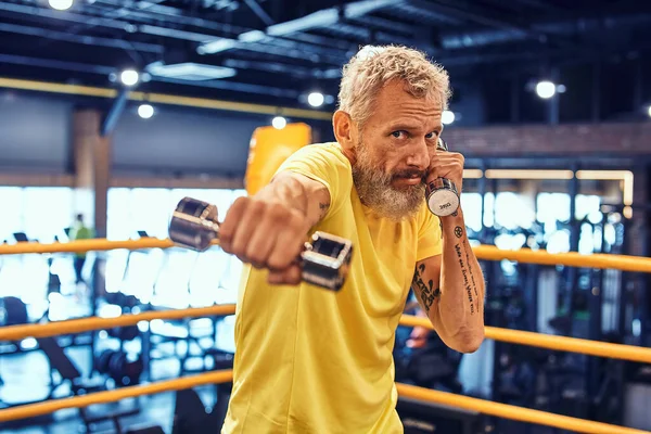 Homem de meia idade em pé no ringue e boxe com haltere e olhando para a câmera enquanto exercita ginásio — Fotografia de Stock