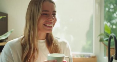 Neşeli bir kadın, huzurlu bir sabahta güneşli bir odada sıcak bir fincan kahvenin keyfini çıkarır.