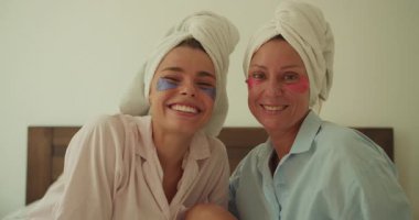 İki mutlu kadın eğlenceli bir spa gününün tadını çıkarıyorlar. Renkli maskelerle gülümsüyorlar ve rahat havlulara sarılıyorlar.