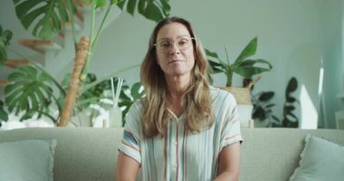 Gözlüklü, güler yüzlü bir kadın koltukta oturur. Etrafı çiçeklerle doludur. Sıcak, davetkar bir atmosfer yayar..