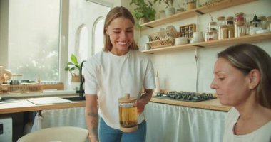 İki kadın, güneş ışığıyla dolu aydınlık ve modern bir mutfakta çay ve kurabiyelerle birlikte güzel bir anın tadını çıkarıyorlar.