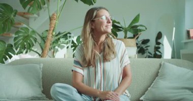 Genç bir kadın şık bir kanepede rahatça oturur, büyüleyici, bitkisel bir yaşam alanında ışıl ışıl gülümser.