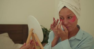 Havluya sarılmış bir kadın, güzellik rutininden zevk alarak aynaya bakarken allık sürer.