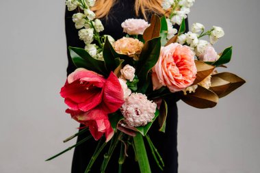 Açık arkaplandaki kızıl saçlı çiçekçinin ellerinde bir buket çiçek. Gül, amilis, östoma, delinyum, manolya yaprakları..