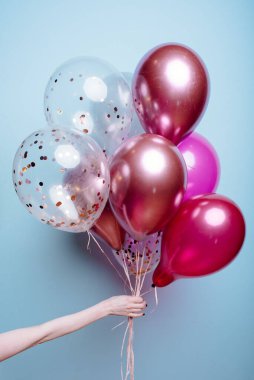 Mavi arka plandaki bir kadının elinde beyaz, pembe, şeffaf helyum balonları var..