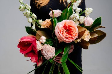 Açık arkaplandaki kızıl saçlı çiçekçinin ellerinde bir buket çiçek. Gül, amilis, östoma, delinyum, manolya yaprakları..