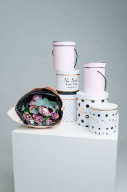 Tozlu pembe pakette bir buket çiçek ve beyaz arka planda beyaz masada şapka hediye kutuları. Gül, sümbül, anemon, okaliptüs.