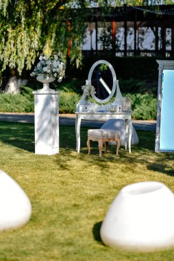Gelin için fotoğraf çekilecek bir yer. Beyaz lüks sandalye, dışarıdaki yeşil çimenlikte bir ayna ve düğün çiçeği aranjmanının önünde duruyor.