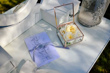 Beyaz çiçeklerin yakın görüntüsü, kutuda alyanslar, içinde bitkiler olan beyaz bir masa, dekoratif bir kafes ve düğün davetiyesi.