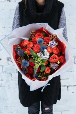 Çiçekler pembe şeffaf pakette. Arka planda örülü süveter giyen çiçekçi kadının ellerinde. Beyaz tuğla duvar..